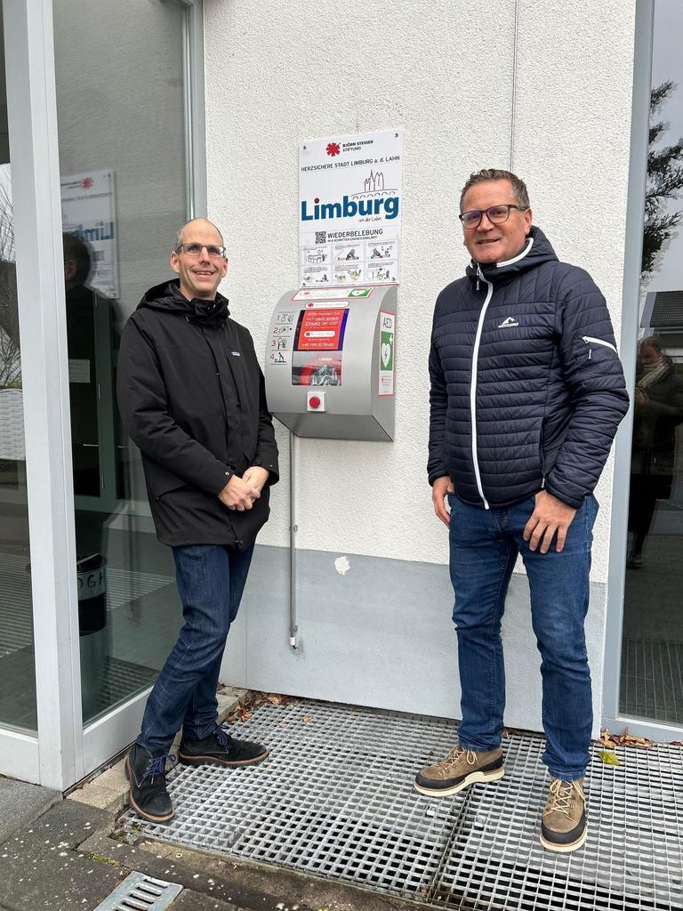  1. Stadtrat Michael Stanke sowie der Leiter des Ordnungsamtes der Stadt Limburg, Michael Wolf vor dem Laien-Defibrillator am Bürgerhaus in Dietkirchen. 